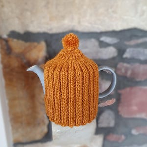 Könnte beinhalten: Eine gestrickte orangefarbene Teekannen-Abdeckung mit einem Bommel oben. Die Abdeckung befindet sich auf einer grauen Teekanne.