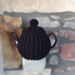 以下が含まれることがあります： 黒いニットのティーコージーが付いた茶色のティーポット。 コージーはリブ編みで、上部に小さなポンポンが付いています。 ティーポットは、湾曲した注ぎ口とハンドルを備えています。