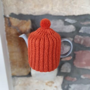 Könnte beinhalten: Ein gestrickter orangefarbener Teekannenwärmer mit einem Bommel oben. Der Wärmer befindet sich auf einer grauen Teekanne.