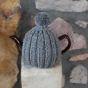 May include: Grey knitted teapot cosy with a pom pom on top. The cosy is on a brown teapot.