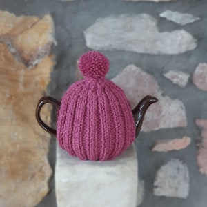May include: A dark brown teapot with a magenta knitted tea cosy. The cosy has a ribbed design and a matching pom-pom. The teapot sits on a white surface against a stone wall.