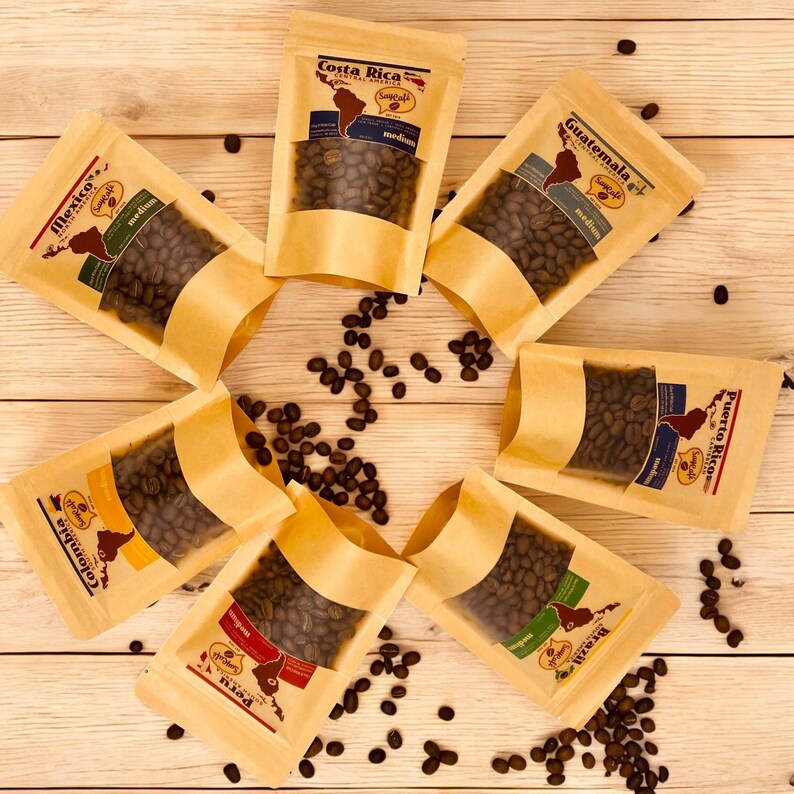 Pu&ograve; includere: Sette sacchetti di carta marrone contenenti chicchi di caff&egrave;. Ogni sacchetto &egrave; etichettato con il nome di un paese di origine, tra cui Costa Rica, Guatemala, Porto Rico, Colombia, Brasile e Messico. I sacchetti sono disposti in cerchio su una superficie di legno.