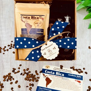Pu&ograve; includere: Una scatola regalo con un nastro blu e bianco a pois, un sacchetto di chicchi di caff&egrave; del Costa Rica e un asciugamano da t&egrave; nero a tema caff&egrave;. La scatola ha un'etichetta "Just Because!" attaccata.