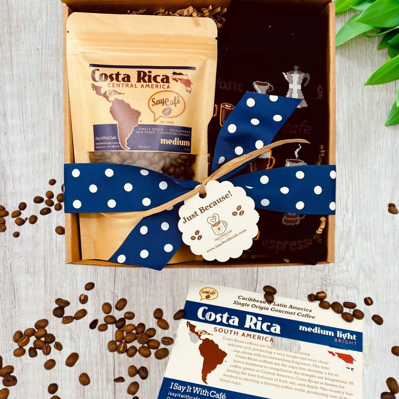 Pu&ograve; includere: Una scatola regalo con un nastro blu e bianco a pois, un sacchetto di chicchi di caff&egrave; del Costa Rica e un asciugamano da t&egrave; nero a tema caff&egrave;. La scatola ha un'etichetta "Just Because!" attaccata.