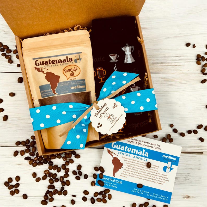 Pu&ograve; includere: Una scatola regalo piena di una busta di caff&egrave; del Guatemala dell'America centrale, un paio di calzini a tema caff&egrave; e un piccolo cartellino che dice "Thinking of You!" La scatola &egrave; legata con un nastro blu con pois bianchi. Ci sono chicchi di caff&egrave; sparsi intorno alla scatola.