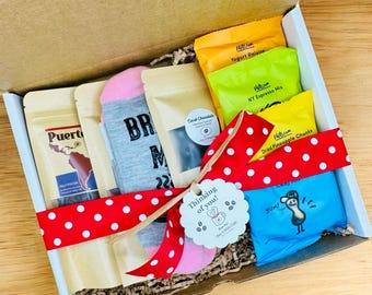 May include: A gift box filled with various items, including a pair of socks with the text "BR" and "M" on them, a bag of coffee, a bag of chocolate, a bag of dried pineapple chunks, a bag of yogurt raisins, a bag of NY espresso mix, and a blue bag with a cartoon bird on it. The box is decorated with a red ribbon with white polka dots and a tag that says "Thinking of you!"