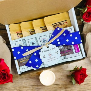 May include: A gift box filled with coffee and tea sachets, tied with a blue and white polka dot ribbon and a "I Love You!" tag. The box is surrounded by red roses and a white candle.