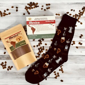 Pu&ograve; includere: Un sacchetto di carta marrone con un'etichetta che dice "Messico Nord America SayCafe Medio" e un calzino marrone a tema caff&egrave;. Il calzino ha uno sfondo nero con tazze da caff&egrave; bianche, chicchi di caff&egrave; e caffettiere. Il calzino &egrave; circondato da chicchi di caff&egrave; su una superficie di legno chiaro.