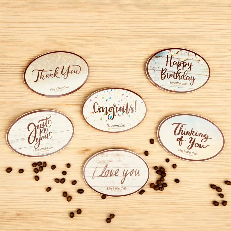 Pu&ograve; includere: Sei etichette regalo ovali con diversi messaggi stampati su di esse. Le etichette sono su una superficie di legno con chicchi di caff&egrave; sparsi intorno. I messaggi sulle etichette sono "Grazie", "Solo per te", "Congratulazioni!", "Buon compleanno", "Pensando a te" e "Ti amo".