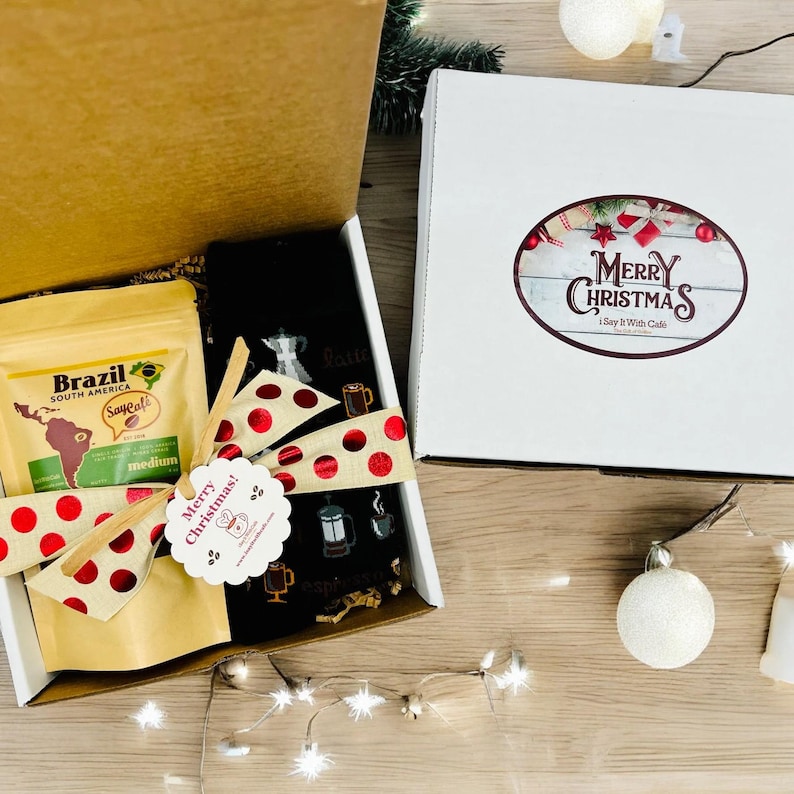 Pu&ograve; includere: Una scatola regalo con un coperchio marrone e un calzino a tema caff&egrave; bianco e nero all'interno. La scatola contiene anche una busta di chicchi di caff&egrave; etichettata "Brasile Sud America" con un nastro a pois rosso e bianco. Un fiocco a pois rosso e bianco &egrave; legato attorno alla scatola con un'etichetta che dice "Buon Natale!"