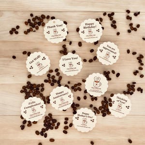 Pu&ograve; includere: Una collezione di 10 etichette regalo rotonde color crema con testo marrone e un design a tazza di caff&egrave;. Le etichette presentano vari messaggi, tra cui "Grazie!", "Buon compleanno!", "Auguri!", "Ti amo!", "Buon anniversario!", "Feliz Cumplea&ntilde;os!", "Sei apprezzato!", "Buona festa della mamma!", "Pensando a te!" e "Say It With Cafe". Le etichette sono sparse su una superficie di legno con chicchi di caff&egrave;.