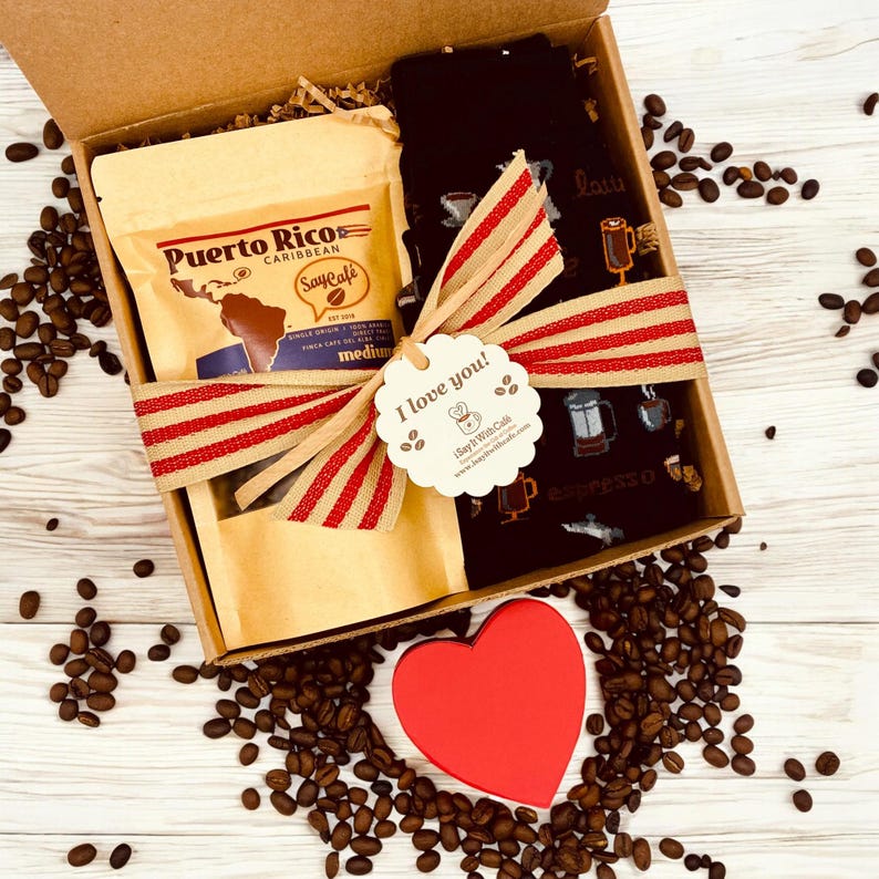 Pu&ograve; includere: Una scatola regalo piena di caff&egrave; e calzini. La scatola &egrave; legata con un nastro a righe rosso e bianco e un'etichetta che dice "Ti amo!" La scatola &egrave; circondata da chicchi di caff&egrave;.