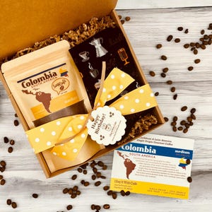 Pu&ograve; includere: Una scatola regalo marrone piena di chicchi di caff&egrave;, una busta di caff&egrave; colombiano e un paio di calzini neri a tema caff&egrave;. La scatola &egrave; legata con un nastro giallo con pois bianchi e un'etichetta che dice "Buon compleanno!"