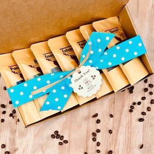 May include: A gift box filled with six small bags of coffee beans, each labeled with a different country of origin. The box is tied with a blue ribbon with white polka dots and a thank you tag.