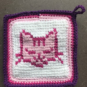 Pode incluir: Um pegador de panela de crochê roxo, rosa e branco com um design de gato. O pegador de panela tem um laço para pendurar.