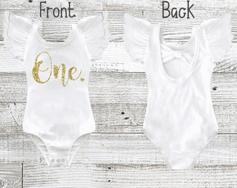 May include: A white bodysuit featuring flutter sleeves and the word One in gold glitter on the front. The back view shows a scoop neckline with a decorative bow detail. The garment includes a snap closure at the bottom for easy access and changing.