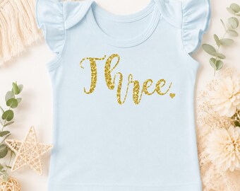 May include: A light blue short-sleeve shirt with ruffled sleeves. The front features the word Three written in a gold glitter script font, followed by a small gold glitter heart. The shirt has a round neckline and a simple, straight hemline.
