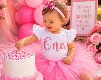 May include: A white bodysuit with ruffled sleeves and the word One in pink glitter, paired with a pink tulle tutu and a matching pink headband with a bow. A white cake with pink frosting rosettes and sprinkles sits on a pink cake stand.