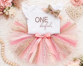 May include: A white short-sleeved bodysuit with ruffled shoulders featuring the rose gold text ONE derful, paired with a multi-layered tulle tutu skirt in shades of pink and gold glitter, and a matching gold glitter bow headband accessory.
