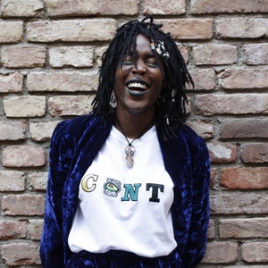 Puede incluir: Una persona con largas rastas negras está riendo. Lleva una camiseta blanca con la palabra "CONT" escrita en letras de colores y una chaqueta de terciopelo azul. La persona está de pie frente a una pared de ladrillo.
