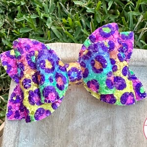 May include: A colorful hair bow with a leopard print design. The bow features a vibrant mix of purple, yellow, green, and blue colors. The bow is made of a textured material and is set against a wooden surface with a grass background.