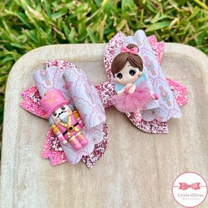 Könnte beinhalten: Zwei dekorative Haarschleifen. Eine Schleife zeigt eine rosa Nussknacker-Soldatenfigur, die andere eine Ballerina-Figur. Beide Schleifen haben eine rosa Glitzerbasis und einen gemusterten Aufsatz mit Nussknacker-Illustrationen.