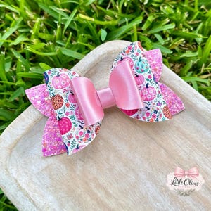 Könnte beinhalten: Eine Haarschleife mit weißem Hintergrund und einem bunten Muster aus rosa, orangefarbenen und türkisfarbenen Kürbissen, Blumen und Blättern. Die Schleife hat mittige Schlaufen aus rosa Satin und Seitenteile aus glitzerndem rosa Material.