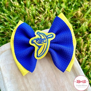 May include: A handmade hair bow featuring a blue and yellow color scheme. The bow has a textured appearance with a yellow border and a blue center. A yellow banana applique with a blue outline is centered on the bow.
