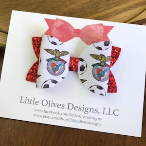 May include: A white and red hair bow featuring a soccer ball pattern and a team emblem. The bow has red glitter accents and is displayed on a white card with the text "Little Olives Designs, LLC".