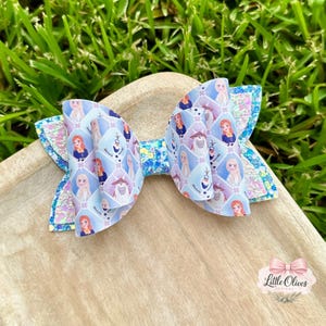 Peut inclure: Un nœud pour cheveux avec un motif en losanges répété représentant des personnages de La Reine des Neiges, dont Anna, Elsa et Olaf. Le nœud est composé de matériaux bleu clair, blancs et bleu pailleté, avec un nœud central et des ailes superposées.
