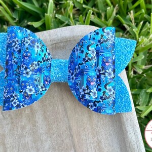 May include: A blue hair bow with a patterned design featuring a cartoon owl and floral accents. The bow has a textured, glittery appearance on the outer edges and a smooth finish in the centre.