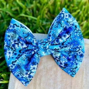 May include: A bright blue hair bow with a floral design. The bow showcases shades of light and dark blue, with white and pink floral details. The bow is crafted from a soft material and is suitable for various events.