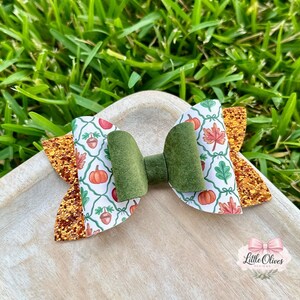 Peut inclure: Un nœud pour cheveux avec un nœud central en feutrine vert olive, entouré de panneaux blancs à motifs répétitifs de citrouilles orange, de glands marron et de feuilles d'automne multicolores, sur une base en tissu pailleté cuivre scintillant.