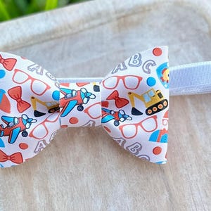 May include: A white bow tie with a pattern of airplanes, eyeglasses, and construction vehicles. The bow tie has a white adjustable strap. The bow tie is on a light-colored wooden surface.