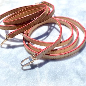 May include: A beige leather strap with a red edge and gold metal clasps. The strap is folded and coiled on a white surface.