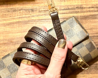 May include: A brown leather strap with gold hardware and stitching. The strap is folded and held in a hand.
