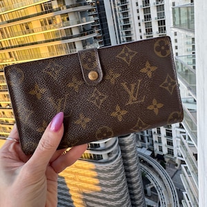 May include: Brown wallet with a gold-colored button closure. The wallet features a repeating pattern of brown floral and logo designs. The wallet is held up against a backdrop of a city skyline.