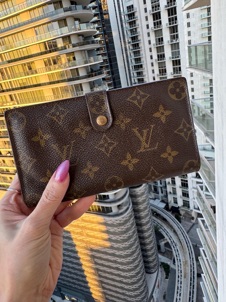 May include: Brown wallet with a gold-colored button closure. The wallet features a repeating pattern of brown floral and logo designs. The wallet is held up against a backdrop of a city skyline.
