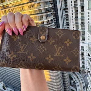 May include: Brown wallet with a repeating pattern of interlocking logos and floral motifs. The wallet has a snap closure with a gold-tone button. The background shows a high-rise building.