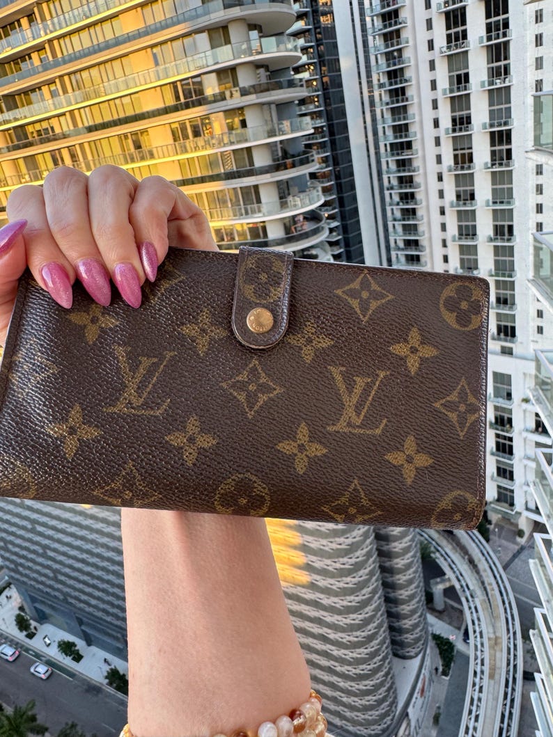 May include: Brown wallet with a repeating pattern of interlocking logos and floral motifs. The wallet has a snap closure with a gold-tone button. The background shows a high-rise building.