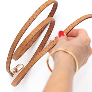 May include: A brown leather strap with gold hardware and a stitched pattern. The strap is coiled and held by a hand.