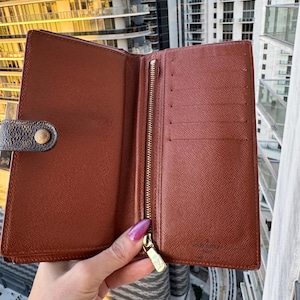 May include: Open brown leather wallet with a gold zipper and snap closure. The interior features multiple card slots and a zippered compartment. The wallet is held open, showcasing its interior design and construction.