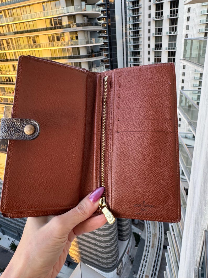 May include: Open brown leather wallet with a gold zipper and snap closure. The interior features multiple card slots and a zippered compartment. The wallet is held open, showcasing its interior design and construction.