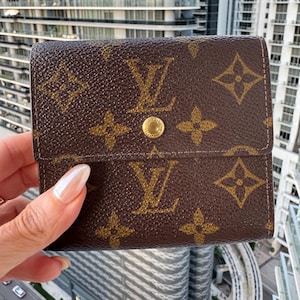 May include: Brown wallet with a repeating pattern of the brand's logo. The wallet has a gold snap closure. The wallet is held in a hand.