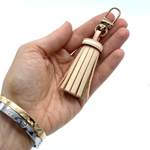 May include: A beige leather tassel keychain with a gold metal clasp. The tassel is attached to a gold metal ring.