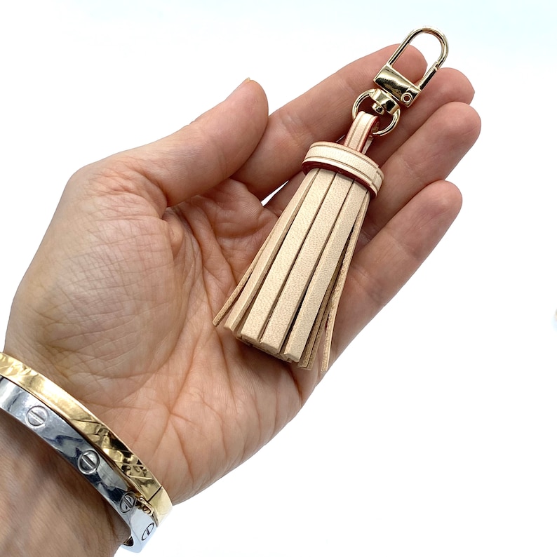 May include: A beige leather tassel keychain with a gold metal clasp. The tassel is attached to a gold metal ring.