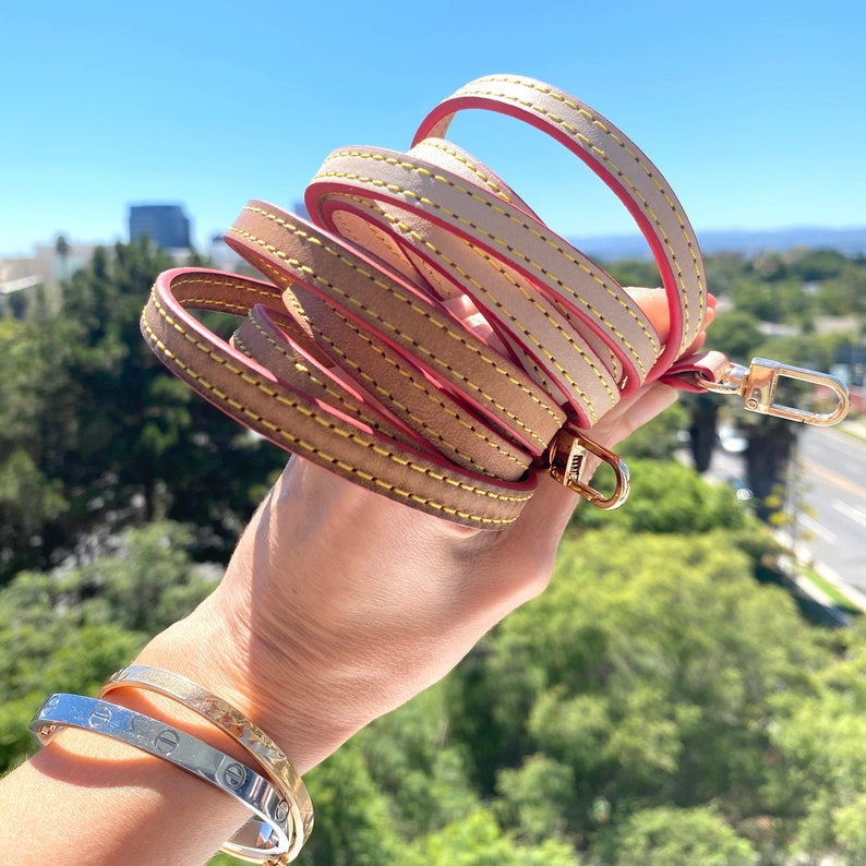 May include: A multi-layered leather strap with a pink stitching detail and gold hardware. The strap is wrapped around a hand.