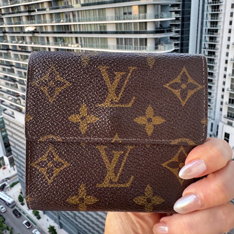 May include: Brown bi-fold wallet with a repeating pattern of gold-colored floral and "LV" monogram. The wallet is made of a textured material and has a snap closure. The background shows a modern building.