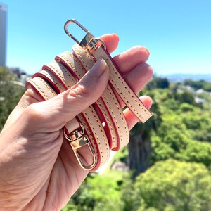 May include: A beige leather strap with red stitching and gold-toned hardware. The strap is folded in half and held in a hand.