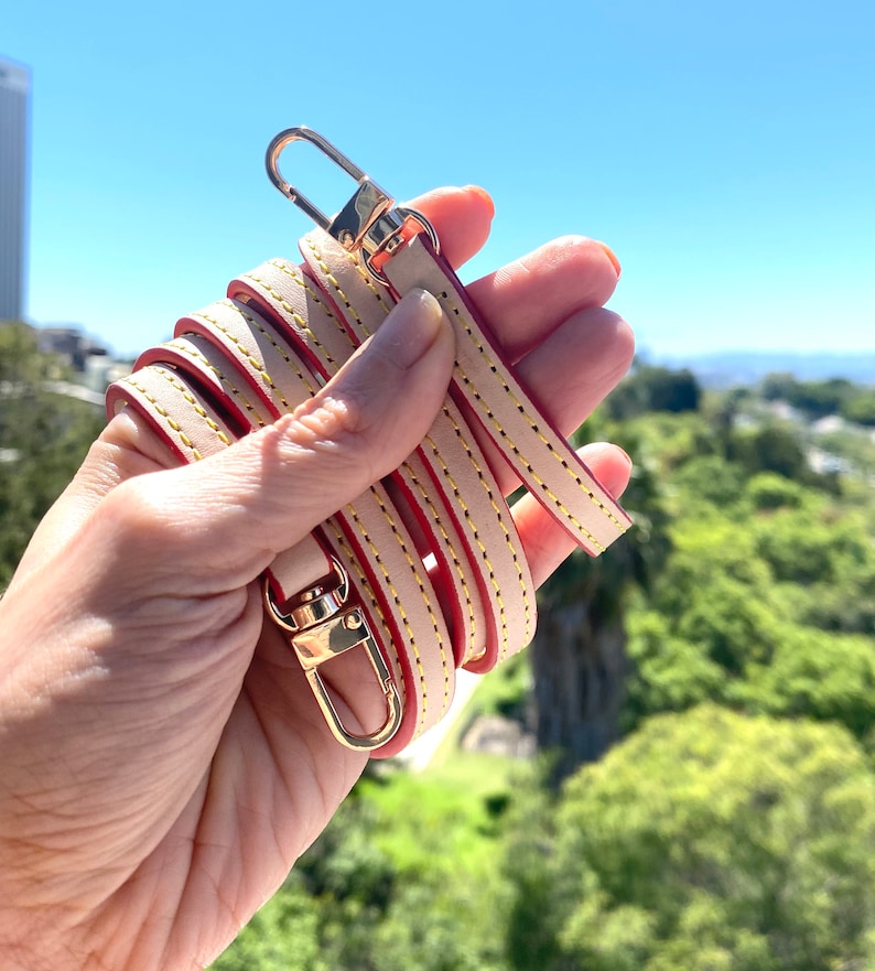 May include: A beige leather strap with red stitching and gold-toned hardware. The strap is folded in half and held in a hand.